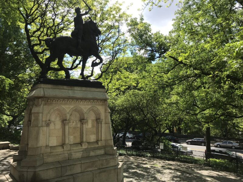 Joan of Arc Statue from the side, Joan of Arc is sitting on a majestic horse. The sky is blue and a bit cloudy and the trees are bright and green.