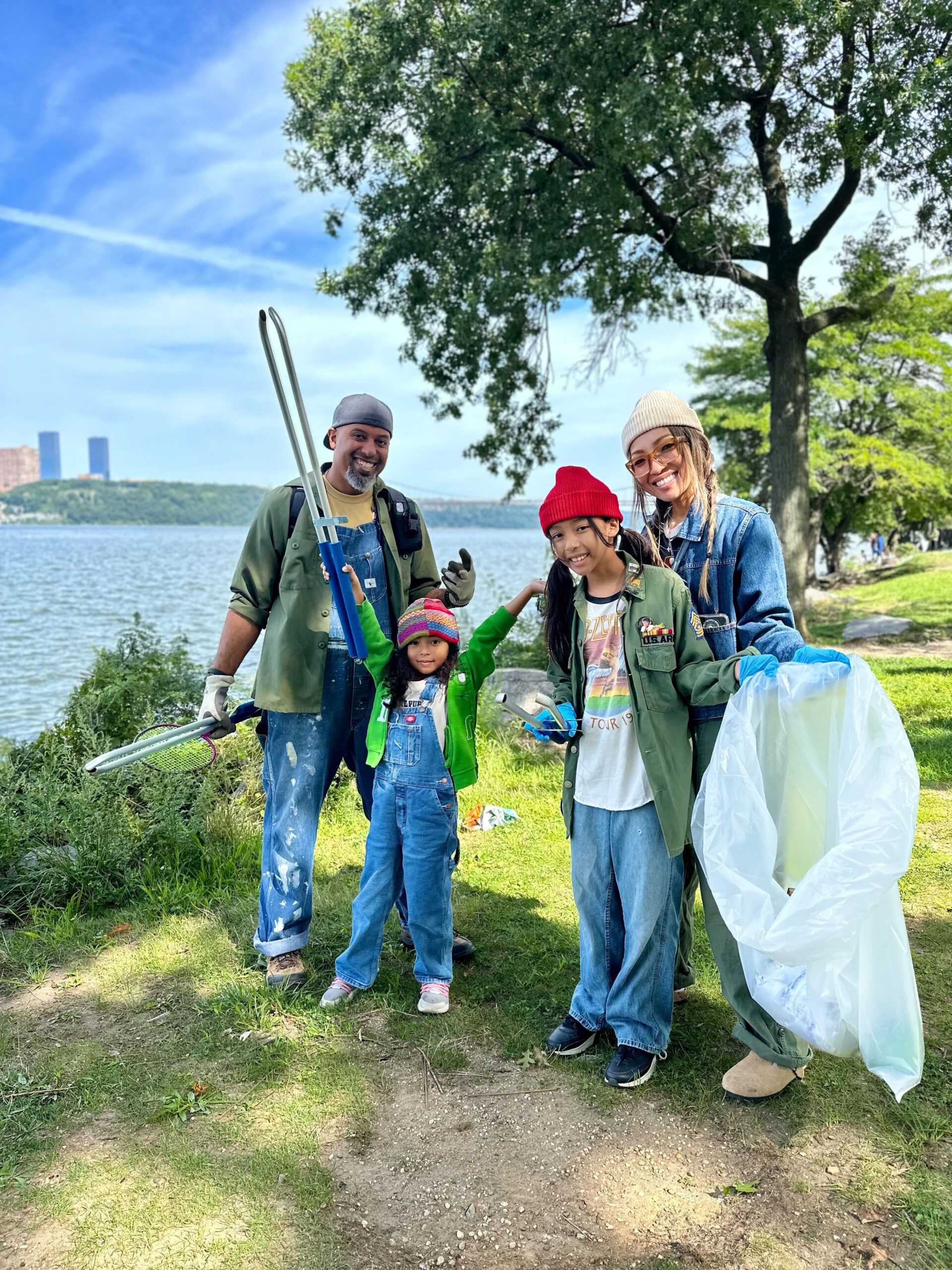 A family of four gathers outdoors by the Hudson River, holding trash bags and litter-picking tools, participating in a cleanup event on a sunny day.