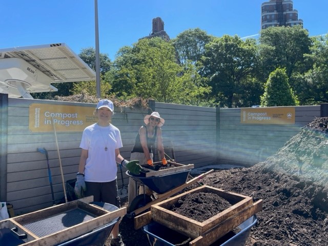 Two volunteers on the left side of the image smile at the camera. They are working with wheelbarrows and sifting compost. To the right is a large pile of compost and signs that say "compost in progress".