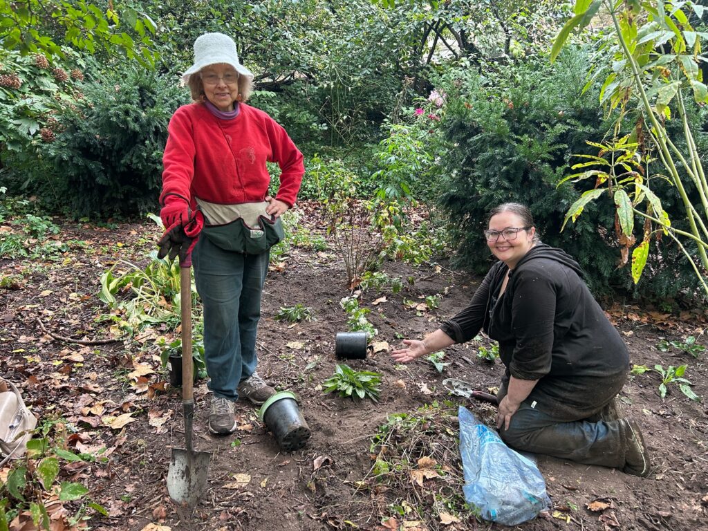 Two volunteers, one standing on the left, one kneeling on the right, in a garden bed in the park. They're surrounded by plant pots and smiling for the camera.
