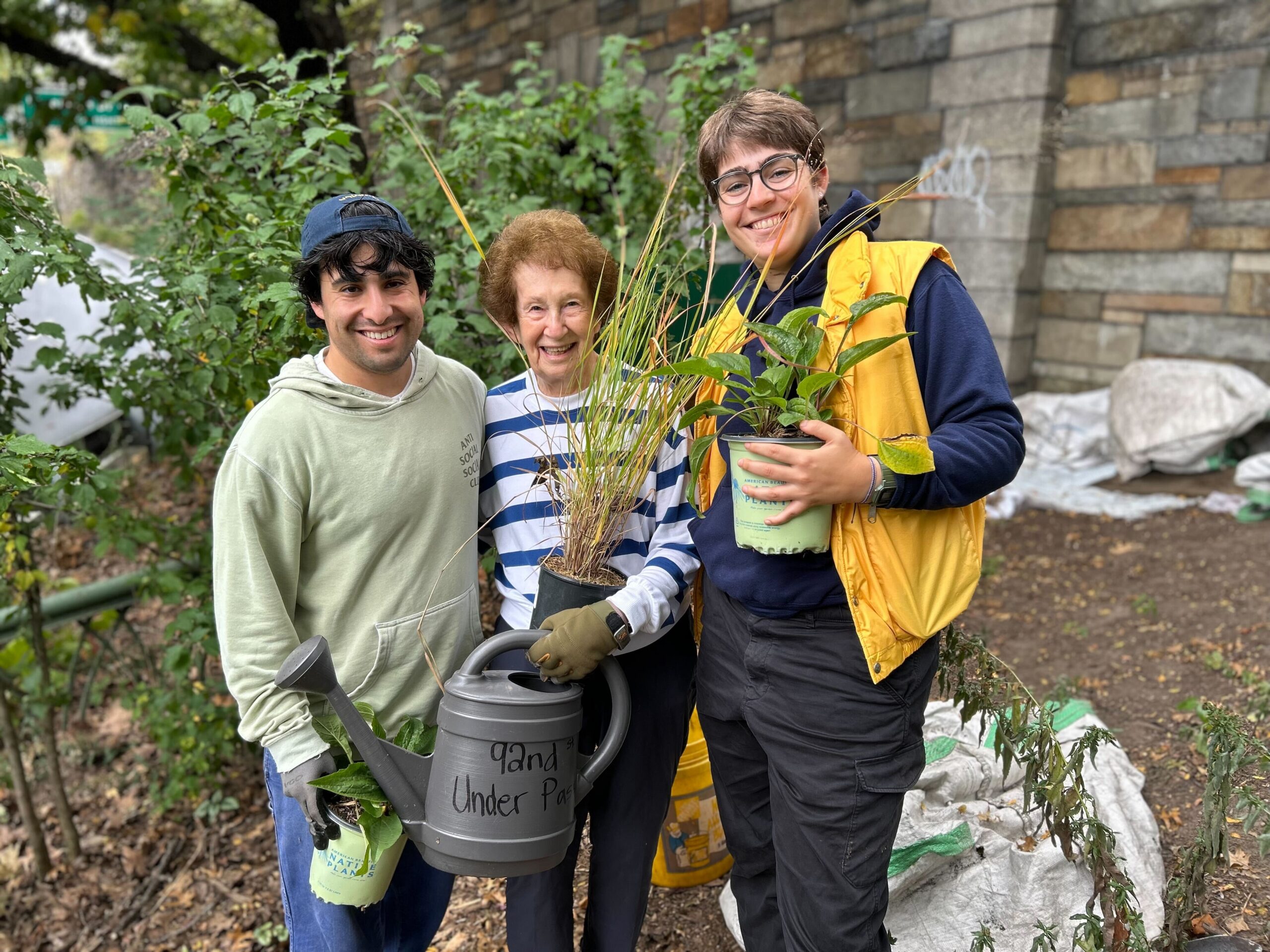 Three people posing for the camera, standing close to each other, holding plant pots and smiling widely. They're standing in a garden bed next to a canvas debris bag.