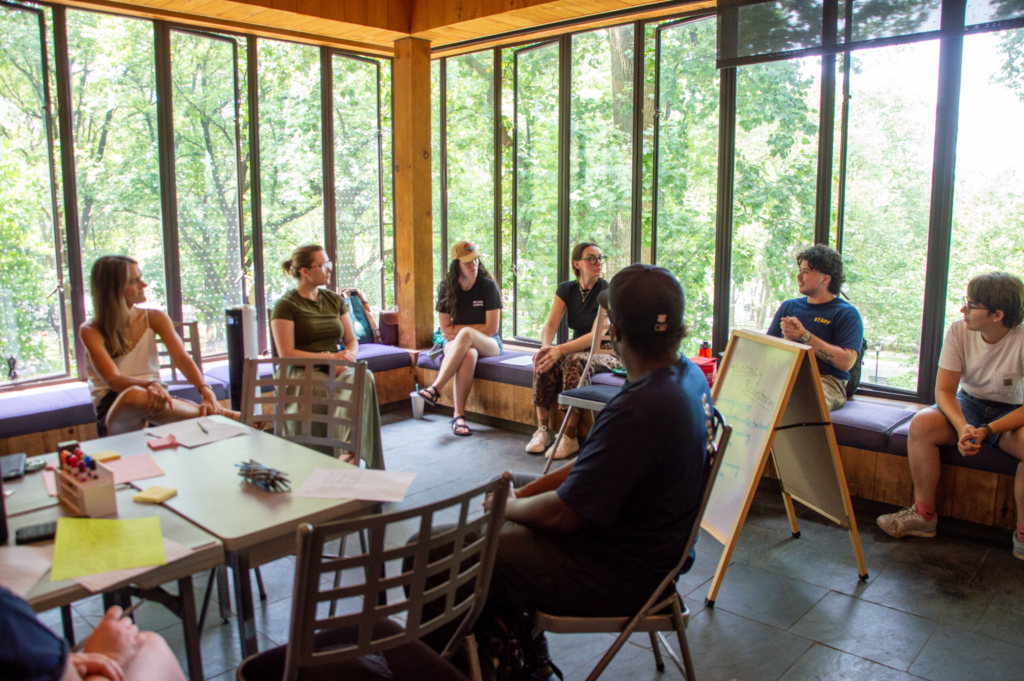 Group of volunteers sitting in chairs and on benches in our Peter Jay Sharp Volunteer House, surrounded by big windows showing green trees outside. There's a whiteboard and the group is discussing Advocacy Club ideas.