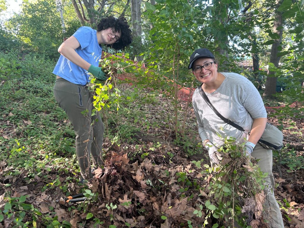 Two volunteers standing on a wooded slope, holding weeds and smiling at the camera.